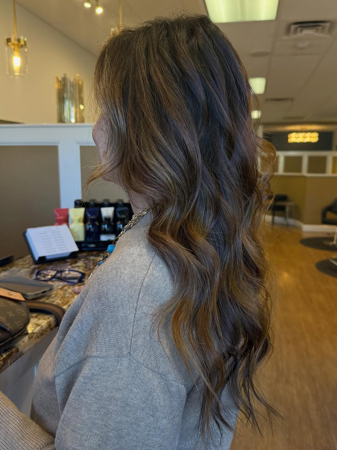 Side view of a woman with long wavy brown hair and subtle blonde highlights in a salon, wearing a gray sweater.