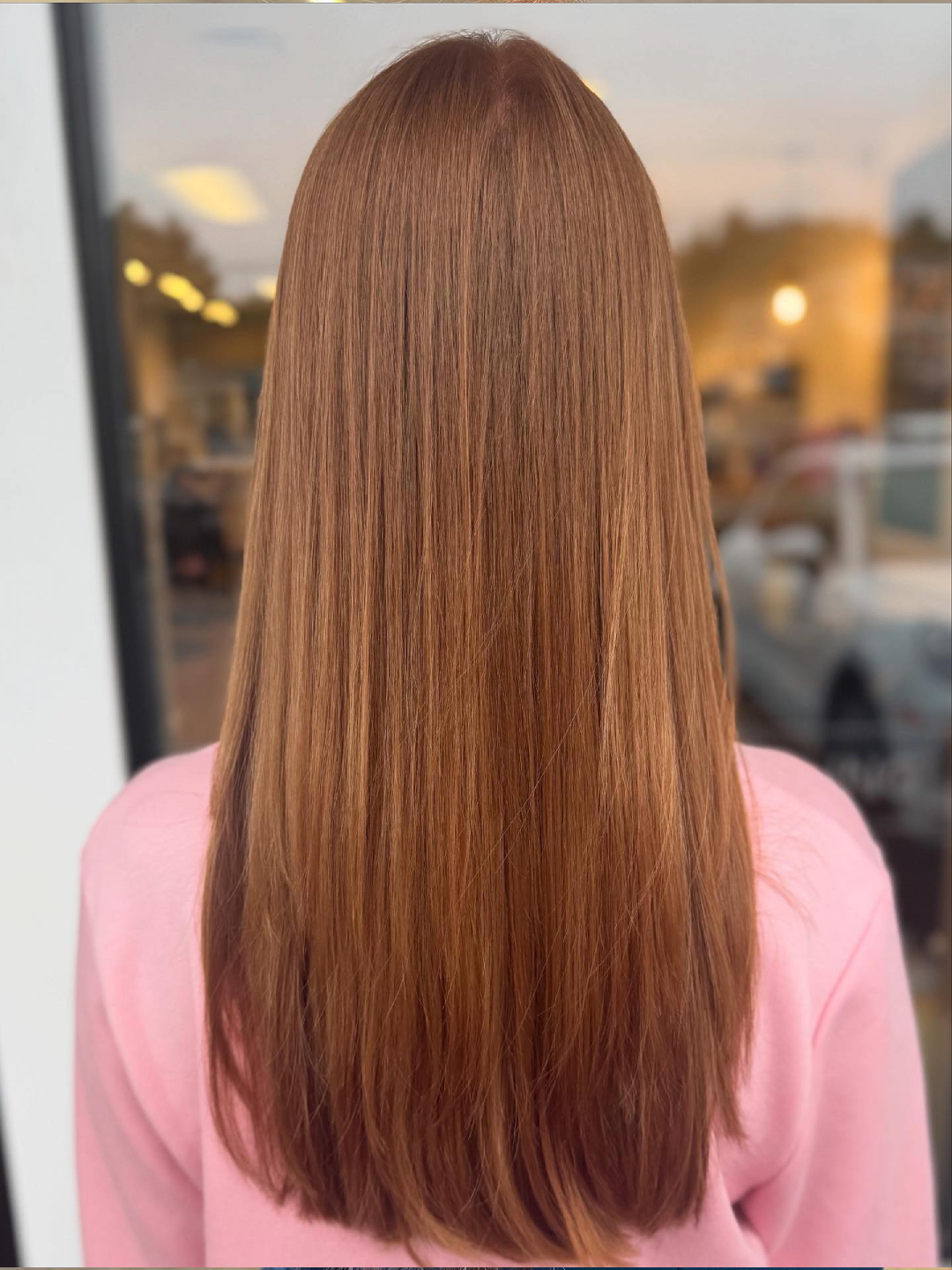 Rear view of a person with long, straight light brown hair, wearing a pink top, standing near a window with soft indoor lighting in the background.