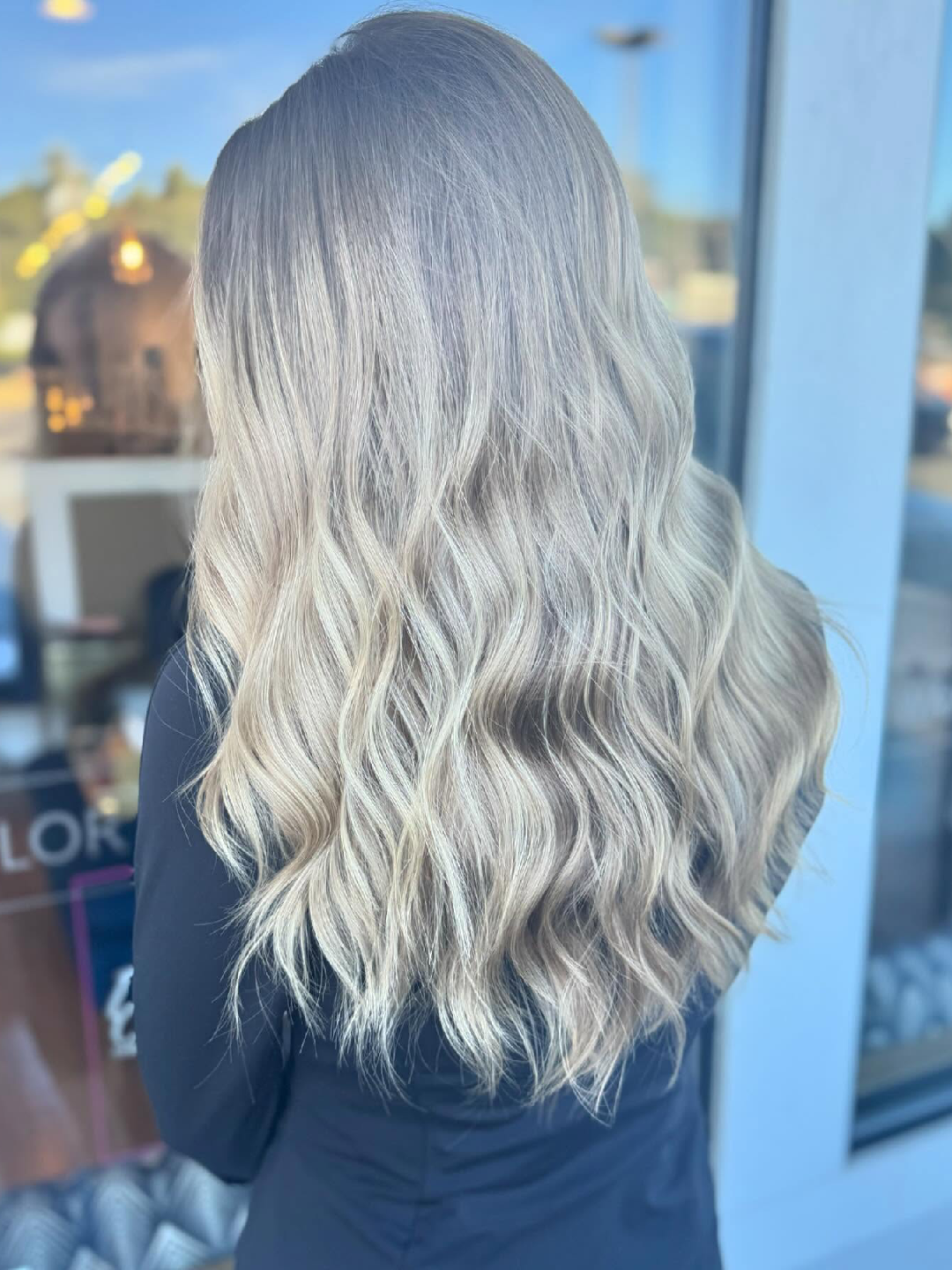 Woman with long wavy blonde balayage hair, viewed from behind outside a glass storefront.