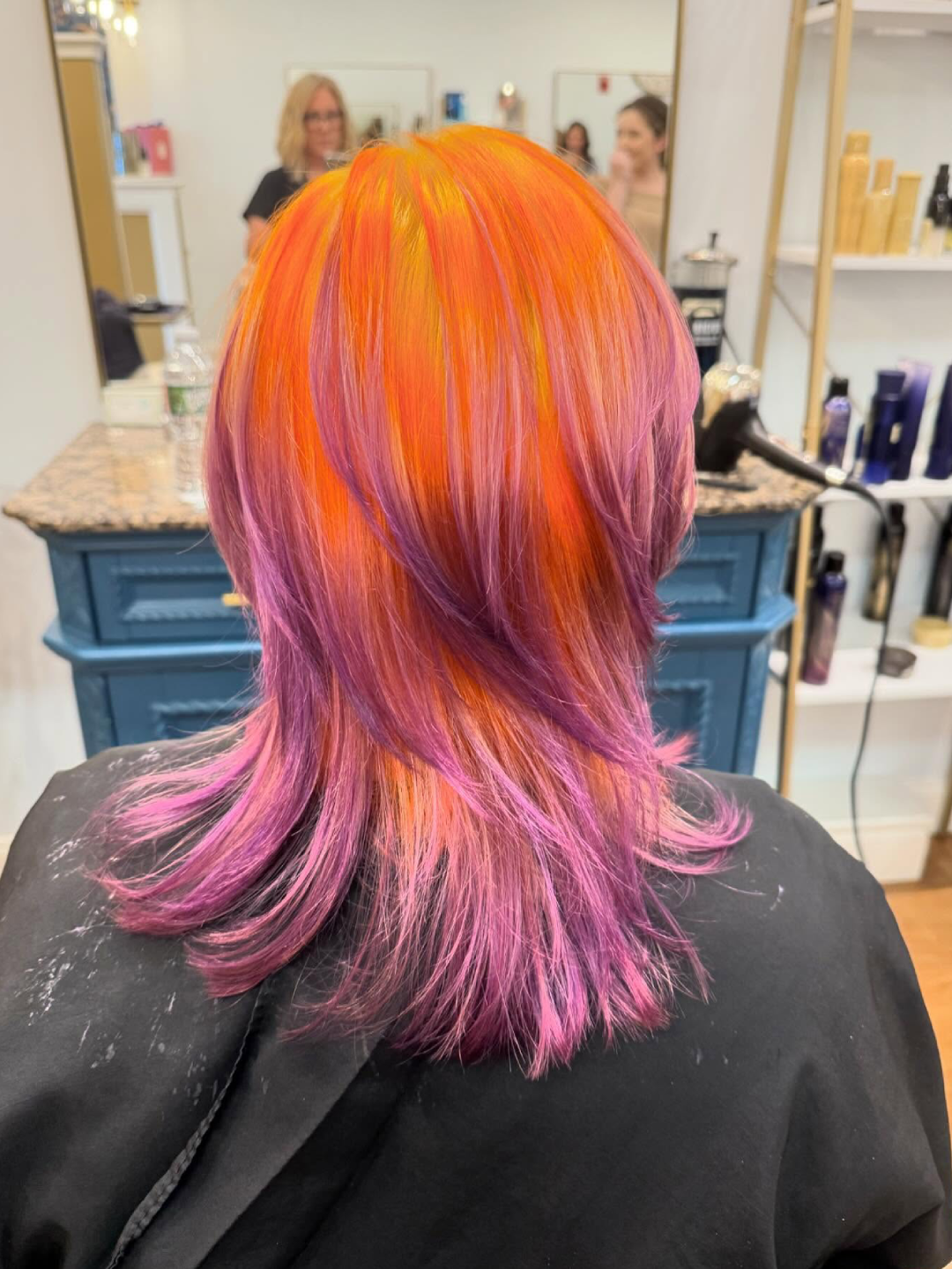 Back of a person’s head with layered bright orange, pink, and purple hair in a salon setting.