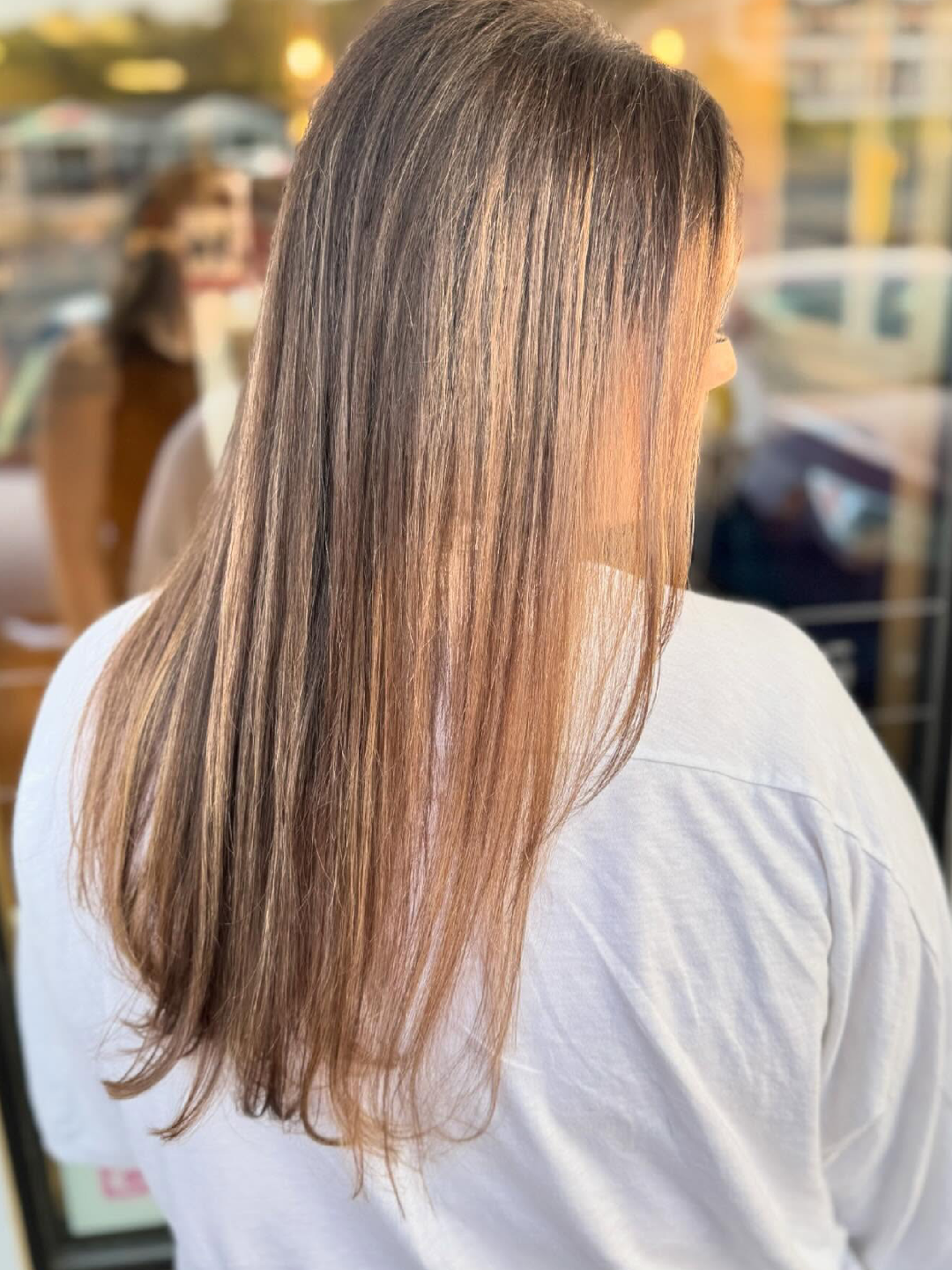 Person with long straight hair, brown with blonde highlights, seen from behind outdoors near a glass storefront.